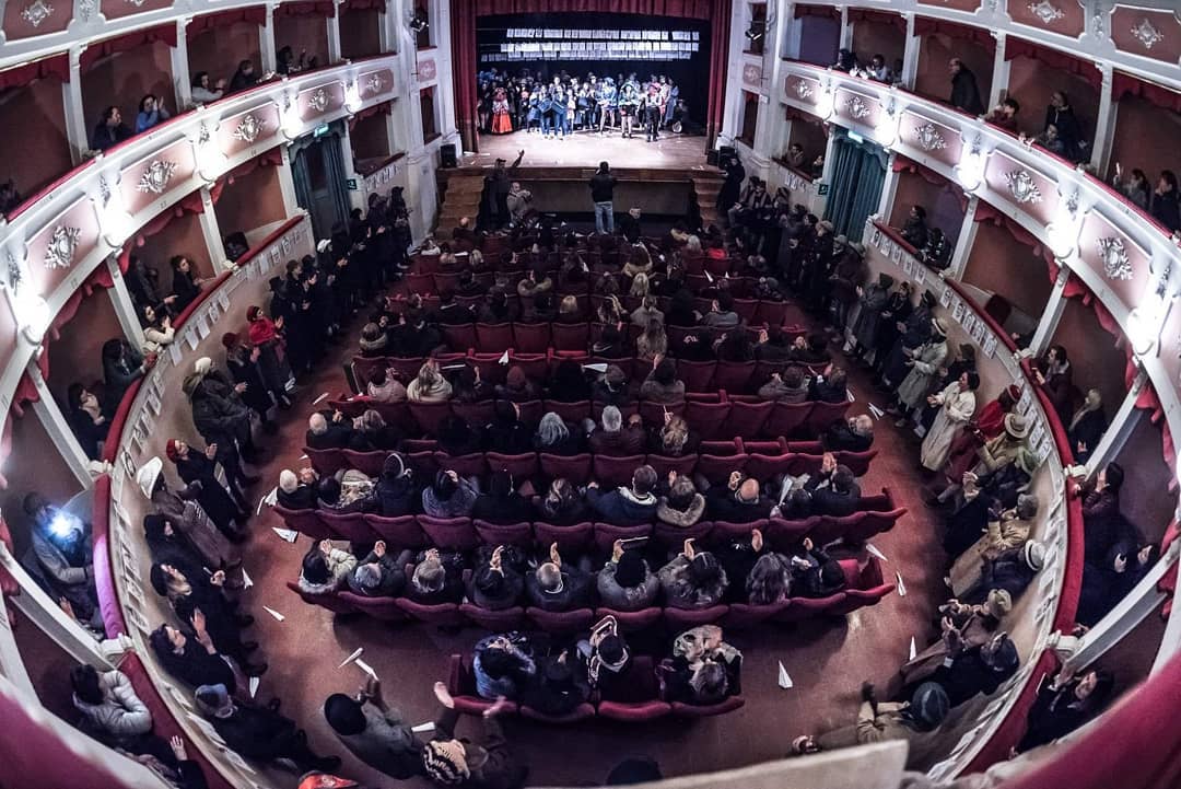 Veduta dall'alto di un teatro storico con la platea gremita di pubblico e i palchetti laterali, illustrazione per il racconto Al Teatro.